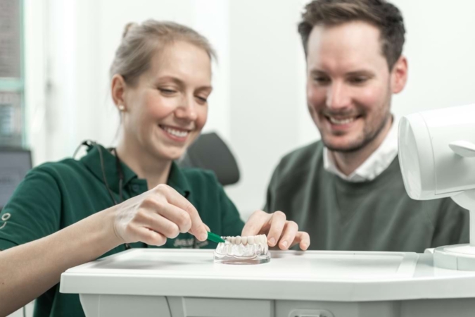 Assistenz zeigt Patient an einem Gebiss, wie man richtig Zähne putzt